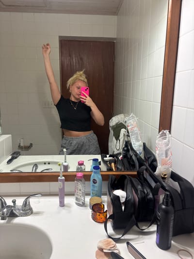 This image depicts a mirror selfie taken by a woman in a bathroom located in Olde Yard Village, British Virgin Islands. The woman, with blonde hair styled in a messy bun, is reflected in a rectangular mirror framed in dark wood. She wears a black short-sleeved crop top and black and white checkered shorts. Her left hand holds a bright pink smartphone, pointed towards the mirror to capture the image. Her right arm is raised above her head, with a clenched fist, in a celebratory or energetic gesture. She wears several rings and a delicate necklace.
The bathroom features white square tiled walls and a dark brown wooden door with paneling visible in the reflection behind her. A white light switch or outlet is mounted on the wall to the left of the door. The white countertop beneath the mirror is cluttered with various personal items. From left to right, these include a black hairbrush, two purple electric toothbrushes, several clear plastic bottles with pink caps, a light blue Nivea After Sun lotion bottle, a small round gold-lidded jar, an amber-colored decorative item, multiple black cosmetic bags, a clear plastic bag containing cotton pads, a black spray bottle, and scattered used cotton pads or makeup sponges. A chrome faucet with two handles and a spout is centered in a white sink basin. A pink hair straightener is partially visible on the far right edge of the countertop. The ceiling reflected in the mirror appears to be white and textured.