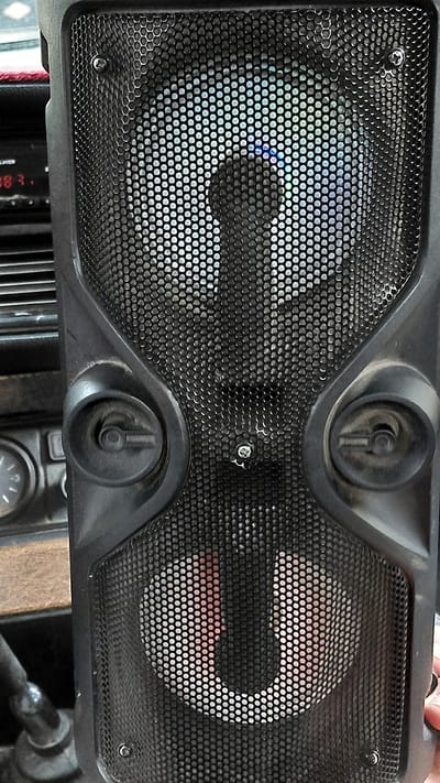 An indoor close-up shot shows a black portable speaker with two circular grilles. The top grille reveals a speaker with colorful LED lights. Below it, two knob-like controls are visible on the speaker's body. The bottom grille also shows a speaker, this one with a reddish hue. To the left of the speaker, part of a vehicle's dashboard is visible, including a gauge with a white needle and numerical markings. The speaker is positioned in a dimly lit environment, suggesting it is not outdoors. The image is cropped, providing limited context about the surrounding area or its specific location within Terek-Say, Kyrgyzstan.
