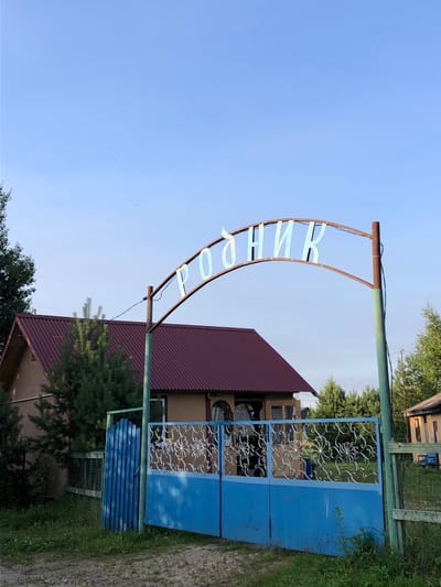 The image shows an arched metal sign with Cyrillic lettering that reads "РОДНИК". The sign is supported by a rusty metal post on the right and another post partially obscured by a building on the left. Below the sign, there is a blue metal gate with decorative scrollwork on the upper half. To the left of the gate is a small blue wooden fence. In the background, a house with a reddish-brown roof is visible, surrounded by greenery. The scene appears to be in a rural or semi-rural setting, possibly a village or dacha community. The sky is clear and blue. Based on the sign's text, which translates to "Spring", and the overall appearance, this location is likely in Makeikha, Russia, possibly marking the entrance to a spring or a property named after one. There are no people visible in the image.