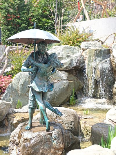 The image is a photograph of a bronze statue depicting a couple dancing under an umbrella. The statue is located in a park or garden setting, likely near a body of water, as evidenced by the presence of rocks, water, and what appears to be a small waterfall in the background. The background also includes trees, plants, and a structure resembling a fence or barrier with people beyond. The statue appears to be the focal point of a park feature or water feature. The image was taken during daylight. The exact location in Верхореченское сельское поселение, Ukraine, is not deducible from the image.