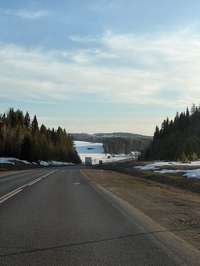 The image depicts a road in a rural landscape near Bol'shaya Sosnova, Russia. The road is asphalt and has double yellow lines marking the center. To the left of the road is a snowy area, likely a field or frozen body of water, with some visible vegetation. Beyond the snowy area is a forested area with evergreen trees. The sky is partly cloudy with a light blue hue. There are no people visible in the image. The photograph appears to have been taken from an elevated position, possibly from an overpass or a higher point along the roadside.