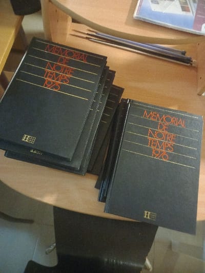 A stack of black books, titled "Memorial De Notre Temps 1975", lies on a light wooden surface. Another stack, bearing the year "1976", is stacked on top. The books feature gold lettering and a small logo at the bottom right. Adjacent to the books are thin, dark objects. The surface appears to be part of a table or shelf. A black chair is positioned nearby.