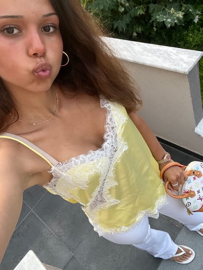 A young woman with long brown hair is taking a selfie outdoors. She is wearing a yellow camisole with white lace trim, white pants, and white flip-flops. She is also wearing a watch on her left wrist and a ring on her right hand. A small white handbag with colorful patterns is held in her right hand. She has a gold necklace on and large gold hoop earrings. Her lips are pursed in a kiss. The background shows some greenery and grey tiled flooring. The lighting suggests it is daytime.