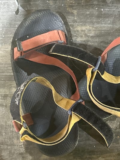 The image displays a close-up, overhead view of two pairs of black sandals with red and yellow straps, resting on a weathered wooden surface. The sandals appear to be outdoor or athletic wear, characterized by their sturdy design and adjustable straps, some of which feature Velcro closures. Small white specks, possibly dust or debris, are scattered across the sandals and the wooden surface. The scene is devoid of any human presence or discernible action. There is no information in the image to suggest a location in Rukoh, Indonesia, or any specific city.