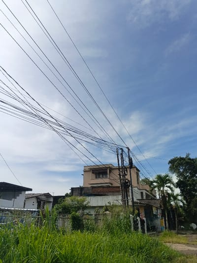 A street scene under a bright blue sky with scattered clouds. Electrical wires crisscross the top of the image, leading to a utility pole. In the foreground, tall green grass dominates the view. Behind the grass are buildings, including a two-story beige house and smaller, more modest structures. Green vegetation surrounds the buildings, and a few palm trees are visible. A road is partially visible on the right.