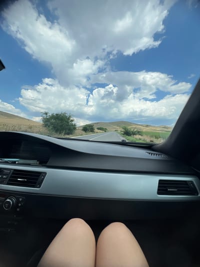 The image shows a view from inside a car, looking out the passenger side window. The scene is a rural landscape with rolling hills covered in dry grass and sparse trees. A road is visible curving through the landscape. The sky is bright blue with large, fluffy white clouds, suggesting a sunny day.

The interior of the car is partially visible on the right side of the frame, showing a textured dashboard with an air vent. A person's legs and knees are also visible in the lower right corner, suggesting they are the passenger.

The overall impression is that of a scenic drive on a clear, sunny day in a rural area. The perspective implies movement, as if the car is traveling along the road. The time of day appears to be daytime, likely late morning or afternoon, given the bright sunlight. The location is identified as Isperihovo, Bulgaria. There is no visible text in the image.
