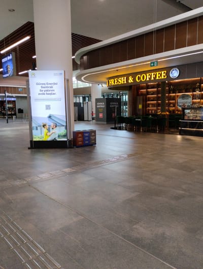The image shows the interior of a modern building, likely an airport or a large transit hub, in Tarsus, Turkey. In the foreground, the floor is paved with large, dark gray tiles, with textured tactile paving strips indicating a pathway. To the left, a large vertical advertisement displays an image of a man and a child looking at solar panels on a house, with text in Turkish promoting solar energy. Next to the advertisement is a white column and a trash receptacle. In the mid-ground, a cafe or coffee shop with "FRESH & COFFEE" prominently displayed in illuminated yellow lettering is visible. The cafe features shelves stocked with items and display cases containing food. Seating areas with dark green chairs are arranged within the cafe. Behind the cafe, more shelves and a patterned ceiling are visible. To the right of the cafe, a "Security Check" sign is posted. The overall environment appears clean, well-lit, and contemporary.