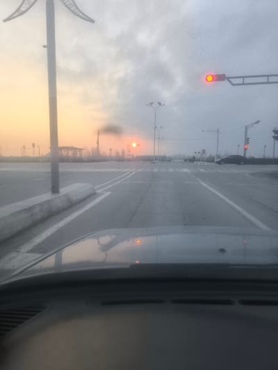The image displays an urban intersection in Pakhtakor, Uzbekistan, at either sunrise or sunset, characterized by hazy, overcast skies. The photograph is taken from inside a vehicle, with the dark dashboard and the top of the vehicle's hood visible in the foreground. The asphalt road surface features white double solid lines and multiple pedestrian crosswalk markings. A concrete barrier lines the left side of the road, near a tall, dark pole with a decorative top. Overhead, a horizontal traffic signal displays a red circular light, and another vertical traffic signal further down the road also appears red. Numerous streetlights with multiple lamp heads are positioned throughout the intersection. On the right side, a dark-colored sedan is stopped near a "STOP" sign. Several indistinct human figures, likely pedestrians, are visible near a distant crosswalk. The horizon, particularly on the left, shows a warm yellowish-orange glow from the low sun, partially obscured by atmospheric haze. A dark industrial structure, possibly a chimney, is visible emitting a plume of dark smoke directly beneath the sun.