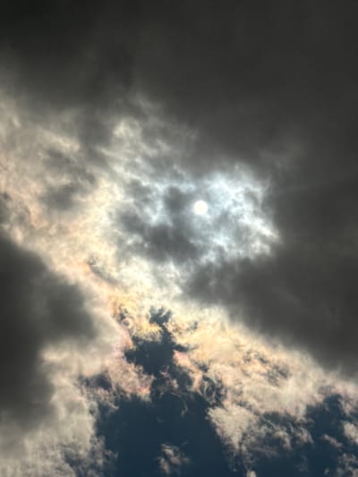 A daytime view of the sky with a sun-like bright spot partially obscured by clouds. The clouds vary in density, ranging from dark gray to lighter white-gray shades. Some clouds show iridescence, with subtle colors like orange, yellow, and pink. The image does not depict Hartavan, Armenia, or any specific location, people, subjects, objects, actions, or interactions.