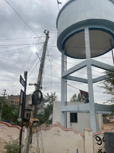 The image depicts a cloudy day in Bethamcherla, India, focusing on a water tank and a utility pole. The water tank, a large, light blue cylindrical structure, is supported by a grey, open-frame concrete base. The tank has a prominent dark blue band around its top and bottom edges. Below the tank, a white building with a window is visible.
To the left of the water tank is a concrete utility pole leaning precariously, heavily laden with a dense network of black electrical wires and cables. Attached to the pole are several items: a street light emitting a bright light, a security camera, and a signpost with two triangular signs, one of which appears to be a directional sign. Near the base of the pole, there is a rusty metal box with a green light, possibly an electrical meter or control unit, and coiled cables.
In the background, a densely packed residential area with multi-story buildings is visible. Some buildings have balconies and tiled roofs. A few trees and foliage are interspersed among the buildings. One building in the background has a flag with an orange symbol. A small, partially visible auto-rickshaw can be seen between two buildings.
In the foreground, a decorative white wall with pink trim lines the bottom of the image. On the wall, there are some inscriptions and large black numerals, including "9". A few green plants are growing in front of the wall. The overall impression is of a densely populated urban or semi-urban environment with visible infrastructure and signs of daily life.