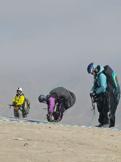 The image depicts three paragliders preparing for takeoff in Alto Hospicio, Chile. The foreground shows the edge of a concrete launching pad painted with blue and white markings. The backdrop features a hazy, overcast sky and distant, blurred mountains. The paragliders are arranged vertically in the frame. The top paraglider is wearing a black and teal suit and a multi-colored helmet. The middle paraglider is wearing a black and purple suit and a dark helmet. The bottom paraglider is wearing a yellow jacket and a white helmet. All are in a pre-flight position, bent over and focused on their equipment.