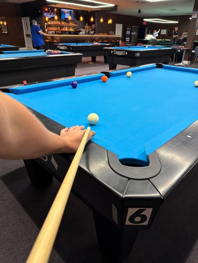 The image depicts the interior of a dimly lit pool hall. The foreground is dominated by a close-up view of a person's right hand gripping a pool cue, poised to strike a white cue ball on a blue felt pool table. Other pool balls, including a purple striped ball and an orange ball, are visible on the table. In the background, several other pool tables are set up. Patrons can be seen in the distance, including a person in a blue shirt standing near a bar and others engaged in playing pool. The environment appears to be a typical entertainment venue, possibly a bar or a dedicated pool hall, with lighting fixtures and a bar counter visible. The location is identified as Palatine, United States of America.