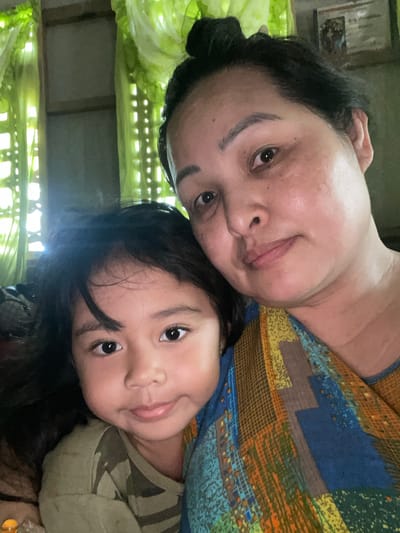 The image depicts a selfie taken indoors, featuring an adult and a child, likely a parent and child. The adult, positioned in the upper portion of the frame, has dark hair pulled back, and is wearing a patterned top with blue, yellow, and brown hues. The child, located beneath the adult, has dark hair and is wearing a green camo-patterned shirt. The background suggests a room with green curtains or foliage visible through window bars or a patterned screen. There is no explicit evidence within the image to confirm the location is in Hijo, Philippines.