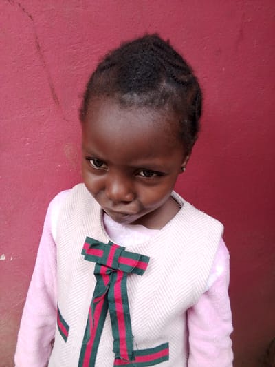 A young Black girl, with braided hair and a small silver earring in her left ear, is standing in front of a red wall. She is wearing a pink long-sleeved shirt underneath a light pink tweed vest that has two green and red striped ribbons tied in a bow on the front. The wall has some scuff marks and a general texture of rough plaster. The girl is looking down and to the left with a slightly pursed expression. The lighting suggests it is daytime, possibly outdoors. The scene is likely in a residential area in Mbar, Nigeria.