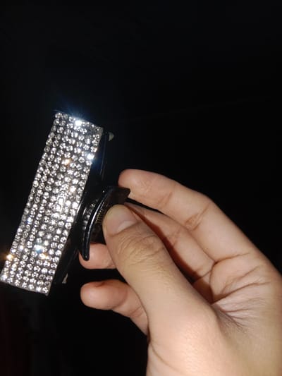 This image, captured in Lam Ujong, Indonesia, presents a close-up view of a human hand holding a black plastic hair claw clip. The top surface of the clip is extensively adorned with multiple rows of small, clear, faceted rhinestones, which brightly reflect light, creating distinct glares. The hand, primarily showing the thumb and a portion of the palm and fingers, is actively gripping the clip, with the thumb positioned to engage the clip's spring-loaded opening mechanism. The background is uniformly dark black, suggesting a low-light environment or an intentionally obscured setting.