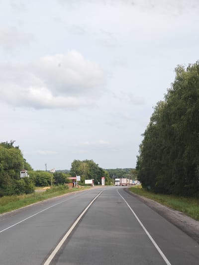The image shows a straight, paved road in a rural setting. The road has a white dividing line and a white line on the right edge. On the left side of the road, there is a grassy verge with trees and a small white structure on stilts, possibly a guardhouse or observation post. Further in the background on the left, there are more trees and a distant building with a golden dome, resembling a church. On the right side of the road, there is a dense line of green trees. In the distance, along the road, several trucks are visible traveling in the same direction. The sky is overcast with light clouds. The scene appears to be located in the village of Липлейка, Russia, given the presence of what seems to be a typical rural Russian landscape with a church and road infrastructure. There are no people visible in the image. The overall impression is one of a quiet, ordinary day on a country road.