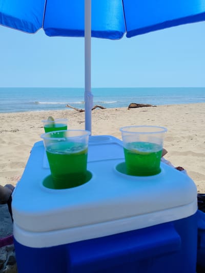 This image depicts a serene beach scene at Novo Oriahovo, Bulgaria. In the foreground, a blue and white cooler is positioned on the sand, with two clear plastic cups containing a vibrant green liquid and lemon slices placed in the cooler's built-in cup holders. A large blue beach umbrella is partially visible, providing shade. The background showcases the calm, blue sea meeting the sandy shore under a clear sky. The beach appears sparsely populated, with a few pieces of driftwood scattered on the sand. The overall atmosphere is one of relaxation and enjoyment of a sunny day by the sea.