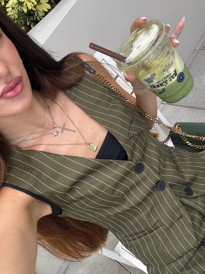 In Hallandale Beach, United States of America, a woman is seen from the chest up, holding a clear plastic cup with a green beverage and a brown straw. The cup has a white lid and features blue and green text that reads "cafe & bakery Honey Veil". She is wearing a green pinstriped vest over a black garment, and multiple gold necklaces adorned with a cross and a small rectangular tag. The background includes a white structure, green foliage, and a portion of a dark green purse with a gold chain strap.