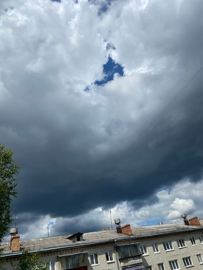 The image shows a view looking upwards towards a dramatic sky filled with a mix of dark, heavy storm clouds and lighter, fluffy white clouds. A small patch of bright blue sky is visible through a gap in the darker clouds. A single power line stretches diagonally across the frame. Below the sky, the roof of a multi-story apartment building is visible, featuring a grey, corrugated roof with several brick chimneys and ventilation pipes. Some trees are visible on the left edge of the frame. The scene appears to be an urban environment, possibly in Beloretsk, Russia, based on the architectural style of the building. There are no people visible in the image.