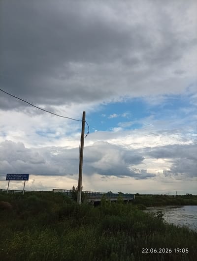 This media file depicts a scenic rural landscape under a dramatic sky. The main subjects include a tranquil body of water on the right, dense green vegetation in the foreground, and a bridge spanning the water in the mid-ground. A wooden pole with a wire extends vertically on the left side of the frame. In the distance, the landscape appears flat with scattered trees and structures, suggesting an open, undeveloped area.
The activity taking place is passive observation of the natural environment, with no people or animals visible. Notable details include the time of day, indicated by the timestamp "22.06.2026 19:05," suggesting it is early evening. The weather appears dynamic, with a mix of dark, heavy clouds and patches of bright blue sky, hinting at changing weather conditions, possibly a storm passing or approaching.
A blue signpost on the left side of the image contains text in both Cyrillic and Latin alphabets. The Cyrillic text reads "ҚАРАҚОБДА өз." and the Latin text below it reads "KARAKOBDA riv.". This identifies the location as the Karakobda River, a tributary of the Ural River in Kazakhstan, near the village of Sarytogay. The overall mood of the image is one of natural beauty, with a touch of atmospheric drama due to the sky.