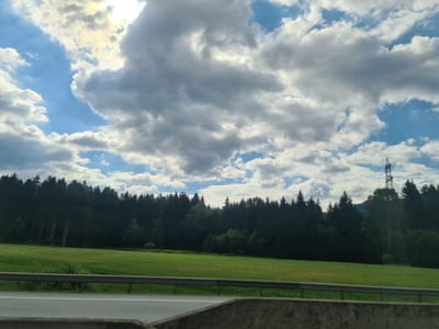 The image depicts a scenic landscape on a partly cloudy day. In the foreground, a concrete barrier separates a road from a lush green field. A metal guardrail runs along the edge of the field. Behind the field, a dense forest of dark green coniferous trees stretches across the horizon. Above the trees, a vast sky is filled with a mix of bright blue patches and large, fluffy white and grey clouds. The sun is visible through some of the clouds, casting a bright glow. In the distance, on the right side of the image, a tall electrical pylon stands against the sky, with power lines stretching across. The overall impression is of a rural or semi-rural environment in Krieglach, Austria.