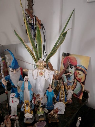 This image, captured in the city of Campo Grande, Brazil, displays an indoor setting, likely an altar or shelf, featuring a diverse collection of religious and decorative figurines. A prominent white statue of Jesus Christ with open arms, adorned with a golden headpiece and a red and gold Sacred Heart emblem on its chest, stands centrally. To its immediate left, a blue-clothed gnome or wizard figurine, complete with a white beard, glasses, and a staff, is positioned. Adjacent to the Jesus statue on the right, a golden, ornate figurine of Our Lady of Aparecida, embellished with small lights or jewels, is visible, flanked by a blue-robed female figurine and a cream-colored angel figurine with golden wings.

Additional smaller figurines are arranged on the dark surface in the foreground, including a white-suited figure with a red tie, a yellow and white-robed figure, and a dark-skinned figure in green attire. A framed print or canvas depicting Krishna playing a flute alongside Radha, who rests her head on his shoulder, leans against the white wall on the right side of the display. A metallic vase or pot containing dried decorative elements is situated to the left of the print.

In the background, a tall, variegated green and yellow snake plant (Sansevieria) ascends against the white wall. Above and behind the plant, a wooden rack or hook system is mounted on the wall, from which various necklaces and keychains, some featuring evil eye charms, hang. Electrical cords are visible behind the plant on the upper right. The display rests on a dark, possibly polished, surface. Pieces of purple amethyst crystals are visible in the bottom right corner of the frame.