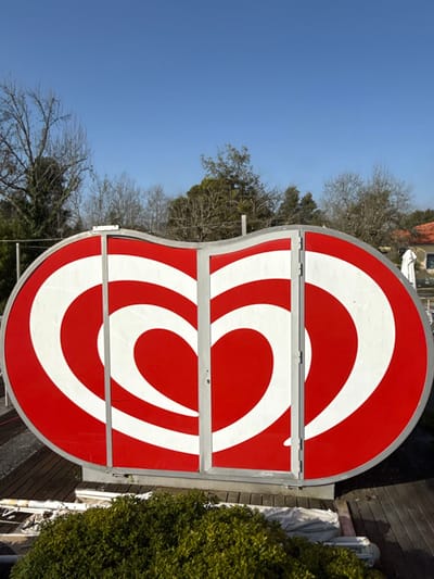 The image depicts an outdoor scene in Mealhada, Portugal, featuring a large, horizontally elongated structure painted with a red and white stylized heart logo. The structure is framed in light grey metal and bisected vertically by a central seam with visible hinges on the right panel, creating two door-like sections. It rests on a dark wooden deck.
In the lower foreground, a dense, manicured green bush occupies the center. To the left of the bush, on the deck, are bundled white tarpaulins and fragmented metallic supports.
The background features a clear blue sky above a line of various trees; some appear deciduous with bare branches, while others are evergreen. To the far right, a section of a building with an orange-tiled roof and a folded white outdoor umbrella is visible. No people or overt actions are present in the scene.
