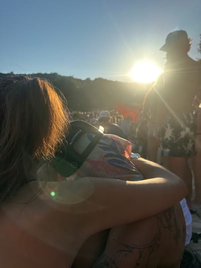 The image depicts an outdoor, crowded event during what appears to be late afternoon or early morning in Tisno, Croatia, given the low, bright sun. In the foreground, two individuals are in a close embrace. The person on the left, partially seen from behind, has long, reddish-brown hair illuminated by sunlight. They wear a black cap with "SUNDAYS" inscribed in white text, and a white, star-like emblem below it. Their arm is draped over the shoulder of the second individual. This second person, also viewed from the back, wears a light-colored, tropical-patterned shirt featuring blue, red, and yellow motifs. Distinct tattoos are visible on their right arm and shoulder area.

Behind these two figures, a dense crowd of people fills the midground, mostly silhouetted or blurred by the strong backlighting. A prominent, bright sun with lens flare dominates the upper right-center, casting intense light across the scene. A red flag or banner is visible among the crowd in the midground-right. The background consists of a clear blue sky above a dark green, forested hillside or treeline, suggesting a natural, possibly coastal or lakeside, setting for the event. The ground beneath the immediate subjects is not clearly discernible but appears to be a natural surface.
