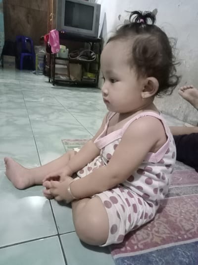 A young child sits on a patterned mat on a tiled floor. The child wears a white outfit with pink polka dots and pink trim. The child has dark hair pulled into a small ponytail on top of their head, and is wearing a gold bracelet and earrings. The background includes a television on a stand, a blue chair, and a cluttered space with various objects. The location is Bungur, Indonesia.