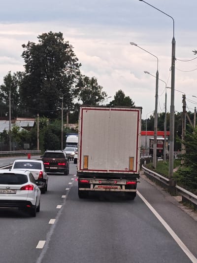 The image depicts a traffic scene on a cloudy day, likely on a highway or main road in Zavidovo, Russia. In the foreground, a white hatchback is visible, followed by a dark SUV, and then a large white semi-trailer truck with red trim. The SUV has a license plate that appears to be Russian. The semi-trailer truck has "Kassbohrer" branding on its rear. In the background, other vehicles and trees are present. Streetlights are also visible along the side of the road. The road is paved and has lane markings. The overall impression is of a typical traffic jam or slow-moving traffic.