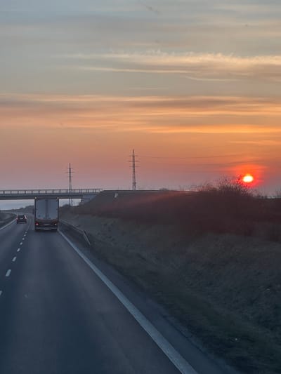 This image presents a highway scene at either sunset or sunrise, located near the city of Slaný, Czechia. The sky exhibits a gradient of colors, transitioning from light blue-grey at the top to pronounced orange and red hues towards the horizon. A distinct, bright red-orange solar disc is visible on the far right, partially obscured by dark, leafless vegetation on an elevated embankment. Thin, horizontally elongated cloud formations are present across the upper sky.
The foreground features a multi-lane asphalt highway with white dashed lane markings separating traffic lanes and a solid white line delineating the shoulder on the right. A dark-colored semi-trailer truck is positioned in the middle lane, heading away from the viewer. Further ahead of the truck, a smaller dark-colored passenger vehicle is also moving in the same direction.
An elevated concrete overpass spans horizontally across the highway on the left side of the frame. Two metal lattice electricity transmission towers are visible in the midground: one situated to the left of the overpass and another positioned to the right, on an elevated piece of terrain beside the highway. The right side of the highway is bordered by a grassy embankment rising gently, behind which is a dense line of dark-toned, deciduous bushes or trees. No human figures are identifiable in the scene.