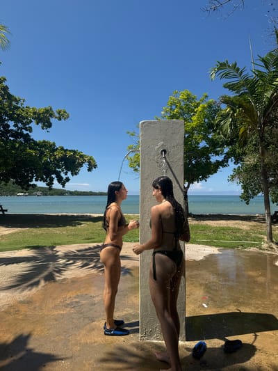 Two females are showering outdoors on a bright, sunny day in Rio Bueno, Jamaica. The scene is set on a concrete paved area adjacent to a grassy lawn that borders a beach and the ocean. A central, light-colored concrete or stone pillar acts as a double-sided outdoor shower structure, with water actively streaming from two spouts.
The female on the left stands in profile, facing right, under a stream of water hitting her head and shoulders. She wears a dark-colored bikini and blue and black water shoes. Her long, dark hair is wet.
The female on the right stands with her back mostly towards the viewer, facing the shower pillar. Water from the second spout streams down her back. She also wears a dark-colored bikini, and her long, dark hair is wet. Two dark items, possibly water shoes or sandals, are visible on the wet concrete near her feet.
The concrete ground is wet from the shower water and displays prominent shadows from overhead foliage, including a palm frond. Beyond the paved area, a manicured green lawn transitions into a sandy beach. The tranquil, blue-green ocean extends into the background, with a distant landmass and a vessel visible on the horizon. Lush green trees and palm trees frame the scene, and the sky above is clear and bright blue.