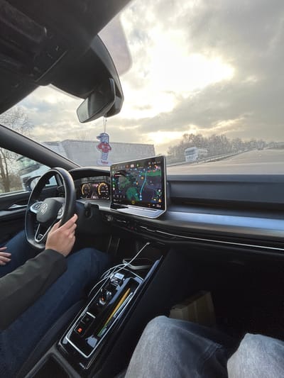 The image captures the interior of a vehicle from the front passenger's viewpoint, located in the vicinity of Hœrdt, France. The driver's hands grip a black, multi-function steering wheel, while their lower torso, clad in blue jeans, is visible. The lower legs of a front passenger, wearing grey jeans, occupy the bottom right frame. The vehicle's dashboard features a digital instrument cluster and a prominent horizontal infotainment display showing a navigation map, current time '15:17', and an outside temperature of '22.0' degrees Celsius. The center console contains a compact gear selector, with a white charging cable connected to a mobile device and a yellow writing instrument. A character-shaped air freshener hangs from the rearview mirror. Outside the windshield, a cloudy sky with bright areas and a multi-lane road are visible. A white tanker truck is observed traveling in the same direction on the right, with additional vehicles in the distance. Bare deciduous trees and a light-colored roadside structure are present.