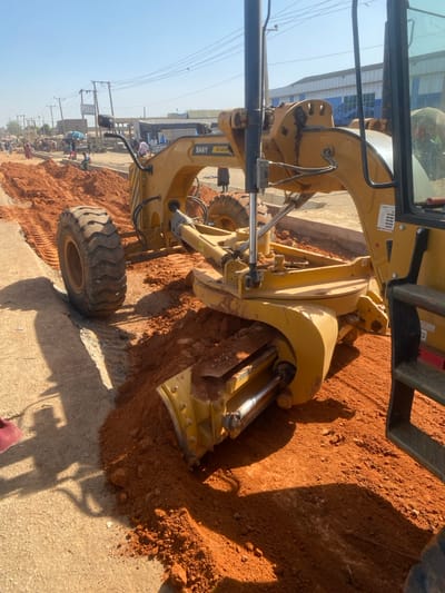 In Rijiyar Lemo, Nigeria, a yellow Sany STG210C-6 motor grader is depicted in operation on a construction site during daylight hours under a clear blue sky. The grader, oriented towards the left, has its central blade engaged with reddish-brown earth, actively excavating and leveling a section of road. Fresh tire tracks are visible behind the grader's large, lugged rear tires. To the immediate left, a compacted or paved surface runs parallel to the disturbed ground. In the background, a busy urban street features multiple pedestrians walking and congregating near various single and multi-story commercial or residential buildings. Some individuals appear to be associated with market stalls, identifiable by umbrellas. Overhead utility lines are strung between wooden poles along the street. The scene is illuminated by strong sunlight, casting distinct shadows, including the lower legs and footwear of an individual visible in the extreme left foreground, positioned close to the grading activity. A larger building with prominent blue-tinted windows stands in the far-right background.