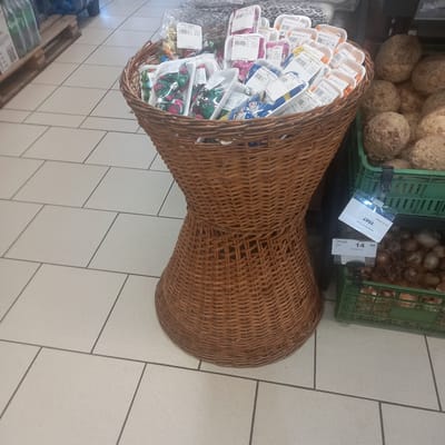In the city of Briceni, Moldova, a store displays a woven wicker basket filled with individually packaged food items, primarily in white plastic containers with printed labels. The items vary in color and shape, with some appearing to be candies or small confections. The basket itself is intricately woven, with a bulbous bottom tapering towards a narrower middle section and then widening again at the top. To the right of the basket, green plastic crates hold root vegetables, likely celery root, and a pile of onions. A white price tag with the number "2507" is attached to one of the crates. Another price tag, bearing the number "14 90" and the word "CRAPA," is visible below the first. The floor is tiled with large, light-colored square tiles. The overall scene suggests a retail environment focused on fresh produce and packaged goods.