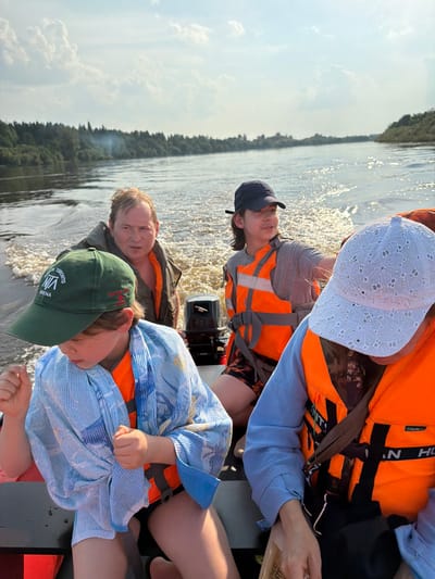 The image depicts a family enjoying a boat ride on a river, possibly in the city of Kalino, Russia, given the presence of dense forests lining the banks and the overall rural landscape. Four individuals are visible in the boat. In the foreground, a child wearing a green baseball cap and a light blue patterned wrap over an orange life vest is looking down. To their right, a person wearing a white, eyelet-patterned baseball cap and an orange life vest is partially visible, with a hint of their face suggesting they are looking forward. Behind them, a young person with a dark blue baseball cap and an orange life vest is looking to the right and gesturing. Further back, a man with light brown hair and an orange life vest is visible, looking towards the camera with a concerned expression. The boat is moving, creating a wake in the water. The sky is partly cloudy, and the sun is shining, casting a glare on the water. A small outboard motor is attached to the back of the boat. The environment appears to be a natural, undeveloped area with trees and water.