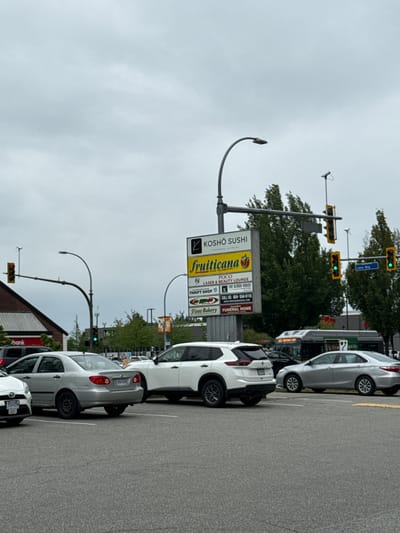 The image depicts an exterior view of a commercial area in Port Coquitlam, Canada, under an overcast sky. The foreground is dominated by asphalt paving, with several vehicles parked and driving. A silver sedan is prominently positioned in the middle ground, facing left. To its right, a white SUV is parked, and further right, another silver sedan is in motion. In the background, a bus with "160 KESTNTOWN LOOP" displayed on its front is visible.

A multi-panelled sign for "KOSHO SUSHI" and "fruiticana" is mounted on a pole. Beneath this, smaller signs advertise various businesses, including "POCO LASER & BEAUTY LOUNGE," "THRIFT SHOP," "The Kebab House," "Poop Bakery," and "FUNERAL HOME," along with contact information. Traffic lights are visible, with one showing a red light and another showing a green light. A street sign reads "Lions Way." To the left of the commercial sign, a building with a red facade has a "bank" sign. Trees and other foliage are present in the background, adding to the urban landscape. The overall scene suggests a typical street view in a suburban commercial district.