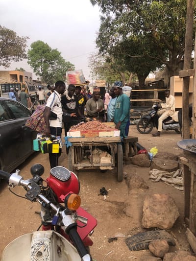 A bustling outdoor market scene unfolds under the shade of lush trees. Several people are gathered around a cart laden with produce. To the right, a red and white motorbike is parked, its handlebars dominating the foreground. In the background, another man sits on a motorcycle. The ground is a mix of dirt, rocks, and debris.