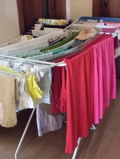 A portable clothes drying rack is positioned indoors, likely in a home. The rack is made of white metal and is filled with various items of clothing that appear to be drying. The clothing includes a mix of colors and patterns, with prominent items being a bright red top and a pink garment in the foreground. Other visible items are a light-colored garment, a yellow piece of fabric, a striped shirt, and some patterned items in the background. The background suggests an indoor environment with wooden doors or cabinets and a wooden floor. There are no people or animals visible in the image. The scene depicts a common household activity of drying laundry. The location of Baden, Austria, is not discernible from the visual information provided in the image.