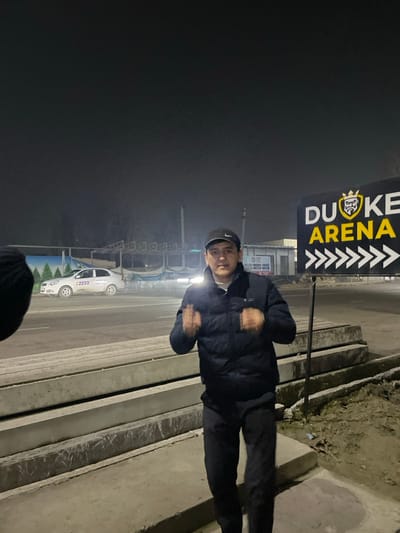A person stands in front of a "Duke Arena" sign with arrows. It appears to be nighttime with a foggy atmosphere. The person is wearing a dark jacket and hat. A car is parked on the street behind them. The ground consists of concrete steps and pavement.