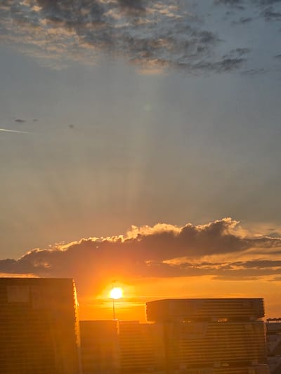 The image captures a sunset over Ansbach, Germany, with a vibrant sky painted in hues of orange, yellow, and blue. The sun, a bright orb, is partially obscured by a layer of clouds, casting a warm glow across the scene. Rays of light emanate from the sun, illuminating the sky and the clouds.
In the foreground, two industrial buildings or structures are silhouetted against the bright sky. These structures appear to be constructed with corrugated metal or a similar material, suggesting a commercial or industrial area of the city. The building on the left is larger and closer to the viewer, while the one on the right is slightly further away.
The sky is dynamic, with scattered clouds adding texture and depth. Some clouds are illuminated by the setting sun, appearing bright orange and yellow, while others are darker and more muted. A faint contrail from an airplane is visible in the upper left portion of the sky, indicating the presence of air traffic.
The overall atmosphere is serene and picturesque, characteristic of a sunset in an urban setting. The image is taken from a slightly elevated perspective, offering a broad view of the sky and the cityscape.