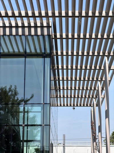 The image shows a modern building with a glass facade and an overhead structure composed of parallel metal slats. The building is located in Rungis, France. The sky is clear and blue. The metal slats cast shadows on the glass, creating a geometric pattern. There are no people visible in the image. The overall scene is clean and orderly, suggesting a corporate or institutional setting.