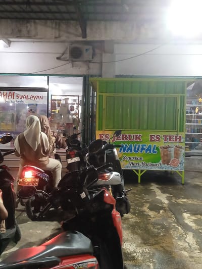 This nighttime image captures an outdoor scene in Sekopek Wetan, Indonesia, in front of a storefront. Two individuals, both wearing head coverings, are seated on a red scooter with its tail light illuminated. They appear to be parked or waiting, possibly outside a store named "AMAI SWALAYAN," which advertises "Lower Price Everyday."
To the right of the scooters is a vibrant green vendor cart, clearly labeled "ES JERUK ES TEH NAUFAL" (Orange Ice Tea Ice Naufal), with the slogans "Segarkan Hari-Harimu!!" (Refresh Your Day!!) and "GULA ASLI" (Original Sugar) below images of iced drinks. Several other scooters and motorcycles are parked in the vicinity, with visible details including a "VARIO TECHNO" and "NAX" brand, and license plates "H 4376 TM 04-27" and "H 4892 AYD 06-28."
The ground is wet, reflecting the artificial lights from above, suggesting recent rain or cleaning. An air conditioning unit is mounted on the wall above the storefront. The overall impression is a bustling, functional urban or suburban environment at night, typical of Indonesian roadside commerce.
