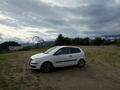 A white, three-door hatchback car is parked on a dirt and grass area. The car is facing the right side of the frame and is angled slightly towards the viewer. Behind the car, a landscape of rolling hills and mountains covered in trees is visible under a cloudy sky. In the mid-ground, a decorative metal heart sculpture is positioned to the left of the car, partially obscuring a wooden fence with prayer flags strung across it. The area appears to be a rural or mountainous region, possibly near Adzhakhy, Abkhazia, given the context of the request. The overall lighting suggests it is daytime, but the clouds create a somewhat overcast atmosphere.