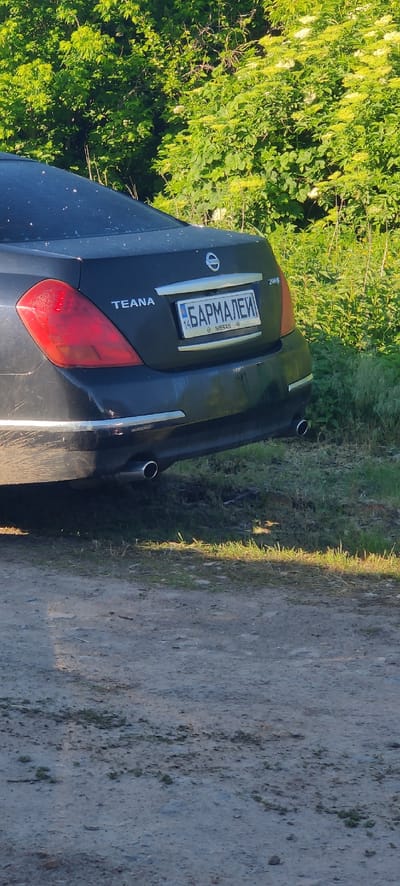The image captures the rear of a dark-colored Nissan Teana sedan, parked on a dirt road or track in Rizunenkove, Ukraine. The car is positioned with dense, lush green foliage, including bushes with light-colored blossoms, to its left. To the right of the car is the unpaved, dusty ground of the road, showing some patches of grass along its edge.
The scene is illuminated by bright daylight, suggesting a clear day. No people are visible.
Notable details include:
- The car's model name "TEANA" is clearly visible on the lower left of the trunk.
- The Nissan logo is on the trunk lid.
- A badge above the license plate appears to say "200R" which might be "2008" or "2009" indicating the car's model year.
- The license plate features Cyrillic text "БАРМАЛЕЙ" (Barmaley), followed by the region code "14" (which corresponds to Cherkasy Oblast in Ukraine), and a small Ukrainian flag emblem with "UA" below it.
- Two exhaust pipes are visible under the rear bumper.
