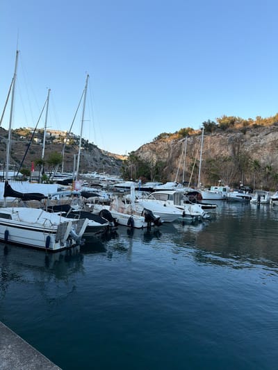 A scenic view of the marina in Almuñécar, Spain, captures a tranquil morning with a clear blue sky. The foreground is dominated by a body of dark blue water, reflecting the surrounding boats and the rocky hills. Several white sailboats and motorboats are docked in the marina, their masts reaching towards the sky. The boats are of various sizes and designs, some covered with dark tarps, others with their sails furled.
Behind the boats, a steep, rocky hillside rises, covered in sparse vegetation and dotted with white buildings that cling to its slopes. The buildings appear to be part of a residential area or a resort, illuminated by the gentle morning sunlight. Palm trees and other greenery are visible on the hillside, adding to the Mediterranean landscape.
The overall impression is one of peacefulness and natural beauty, with the marina serving as a focal point against the dramatic backdrop of the Spanish coastline. No people are visible in the image, and the boats appear to be at rest. The water is calm, with gentle ripples on its surface.