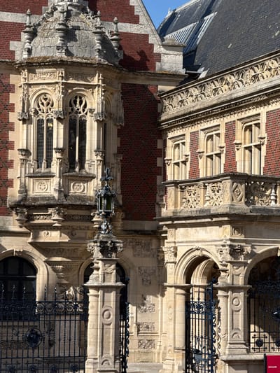 The image captures a detailed view of an ornate, historic building from a low, upward-looking perspective, under bright, clear daylight. The architecture combines striking red brickwork, often laid in patterns, with extensive sections of light-colored, intricately carved stone.
The building exhibits characteristics of Gothic or Neo-Gothic design, featuring elaborate stone tracery around windows, pointed arches, and decorative cornices. A steep, dark slate roof with dormer windows is visible against a clear blue sky in the upper left. The facade is adorned with a variety of sculpted elements, including floral and foliate motifs, geometric patterns, and grotesque figures. One prominent detail is a large, winged creature, possibly a dragon or griffin, extending from the facade, appearing to hold a lantern or escutcheon.
Several windows with dark or stained glass are visible, some rectangular with circular elements, others with more complex curvilinear tracery. Wrought-iron railings with decorative scrollwork and finials protect what appear to be balconies or openings on various levels. Strong shadows cast across the facade emphasize the intricate three-dimensional relief of the carvings and architectural features, indicating a sunny day.
No people are visible, and no specific activity is depicted. A red sign with white text, partially visible behind a wrought-iron gate, clearly displays the word "DISTILLERIE". This sign serves as a significant location cue, strongly suggesting the building's historical or current function. The building's grandeur and detailed craftsmanship dominate the scene.