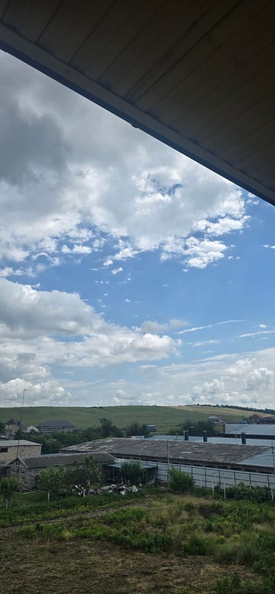 The image captures a panoramic view of the outskirts of Suvorovskaya, Russia, under a bright, partly cloudy sky. The foreground is dominated by a section of wooden roof extending from the top left, casting a shadow that frames the scene. Below the roof, a vast expanse of sky stretches out, filled with puffy white clouds against a clear blue backdrop.

The middle ground reveals a landscape dotted with buildings and greenery. Several low-lying structures, appearing to be agricultural or industrial buildings with corrugated metal roofs, are visible. Some of these have chimneys or ventilation shafts. Adjacent to these structures are areas of dense green vegetation and trees. Beyond the immediate buildings, rolling green hills rise, with scattered trees and a few more distant buildings visible on their slopes. Faint lines of what might be fences or pathways can be observed on the hills.

The background is a soft, hazy horizon line where the green hills meet the sky. The overall impression is one of a rural or semi-rural environment on a day with dynamic cloud formations. No people are clearly discernible in the image. The scene is static, with no apparent actions or interactions taking place. The lighting suggests it is daytime, likely late morning or afternoon, given the brightness of the sky and the contrast of the shadows.