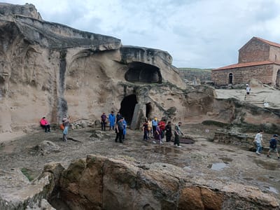 This image captures a group of tourists exploring the ancient cave city of Uplistsikhe, Georgia, under a cloudy sky. The foreground is dominated by rugged, rocky terrain, with scattered pools of water reflecting the overcast sky, indicating recent rainfall. In the midground, a large, carved rock formation with multiple cave entrances is visible. Several individuals are scattered throughout the scene, some appearing to be guides or part of organized tour groups. One person in a bright pink jacket sits on a ledge carved into the rock face. Further to the right, a stone church with a red-tiled roof stands prominently, its architecture typical of Georgian historical churches. The ground around the church is also rocky and uneven, with some stone steps leading up to its entrance. The overall atmosphere suggests a historical and archaeological site being visited by tourists.