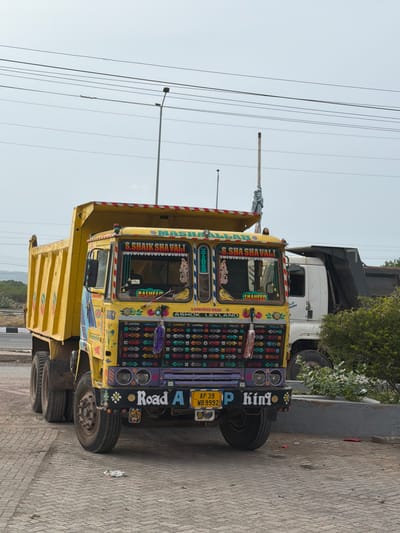 The image shows a yellow and colorful truck parked on a patterned brick surface. The truck is decorated with various colorful designs and text. The license plate reads "AP 39 WB 9992". In the background, another white truck is partially visible, along with some green bushes and a grey wall. The sky is overcast. The location is identified as Thammarajupalle, India. There are no people visible in the image.
